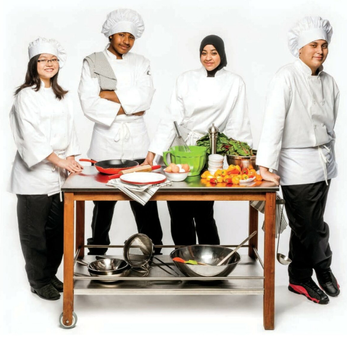 Four teenage students standing at a table. The students are all dressed in Chef attire.