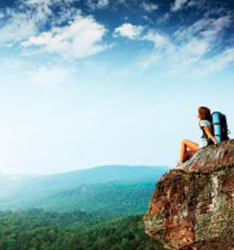 A photo of a traveller sitting on a cliff ledge overlooking an expansive forest. The sky above is blue and white, smattered with clouds.