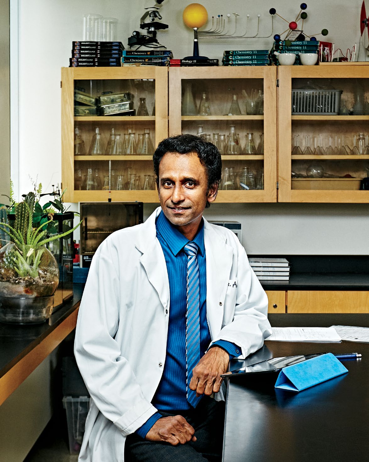 A photo of Gabriel Ayyavoo, Ontario Certified Teacher, seated at a desk in a science class. Gabriel is dressed in white lab technician attire.