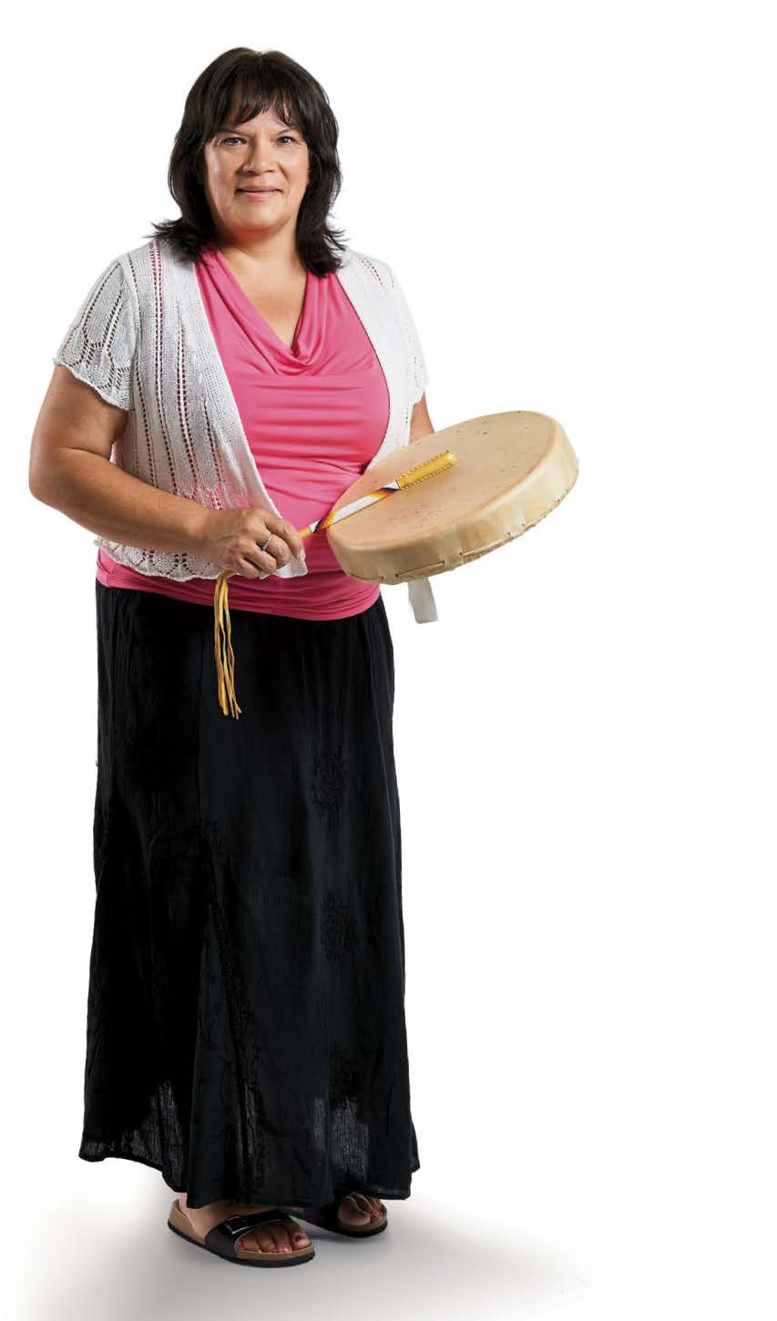 Debbie Debassige standing. Debbie is holding a drum in one hand and an ornate drumstick in the other. 