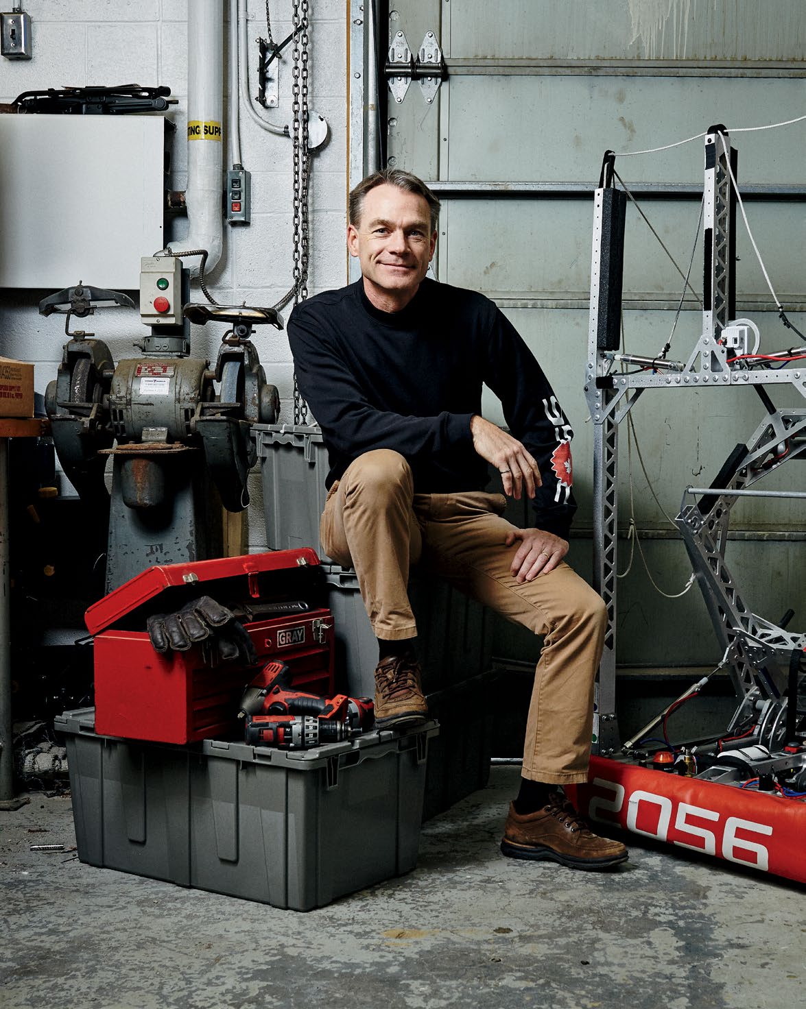 Photo of Stanley Hunter seated on a storage container with one foot resting on another storage container. There is a mechanical contraption as large as Stanley to the right. 