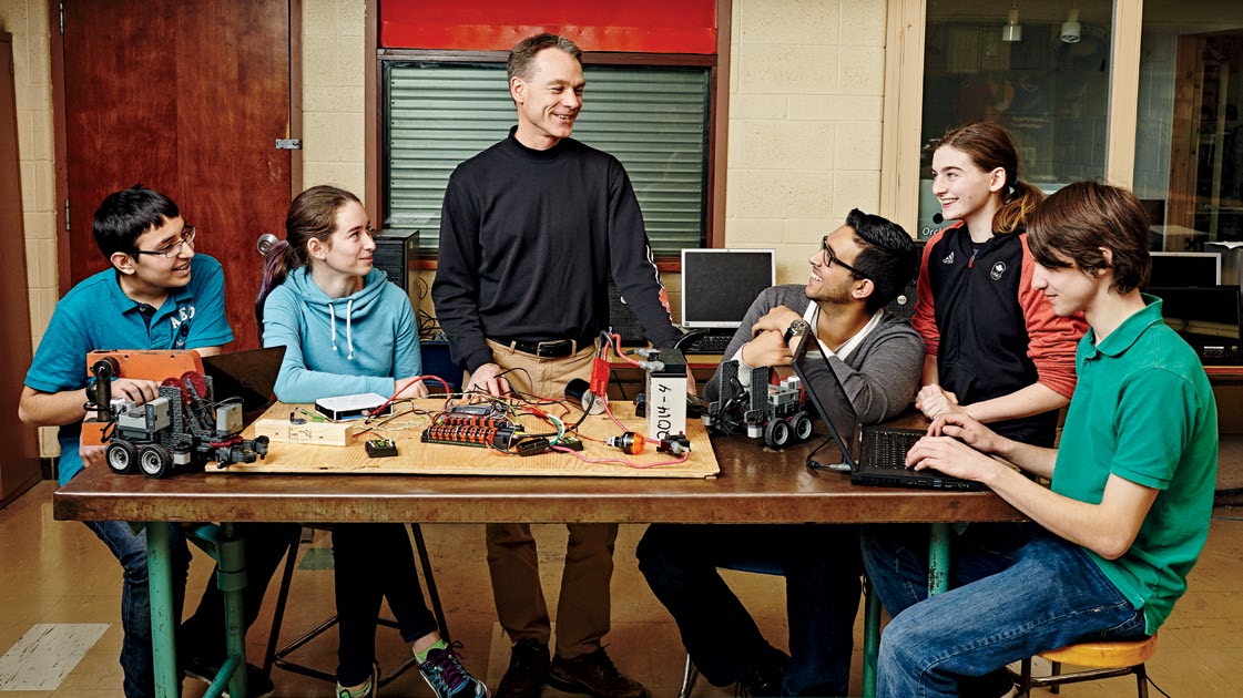 Photo of Stanley Hunter standing at a work table. There are a variety of electronics components on the table. Four students are seated around Stanley. One of the students works on a laptop while the others look at Stanley.