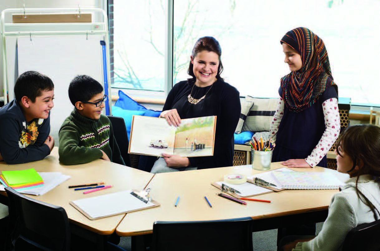 Tracey Tinley holds a book open while seated at a desk. Tracey is surrounded by four young students who are looking at the book.