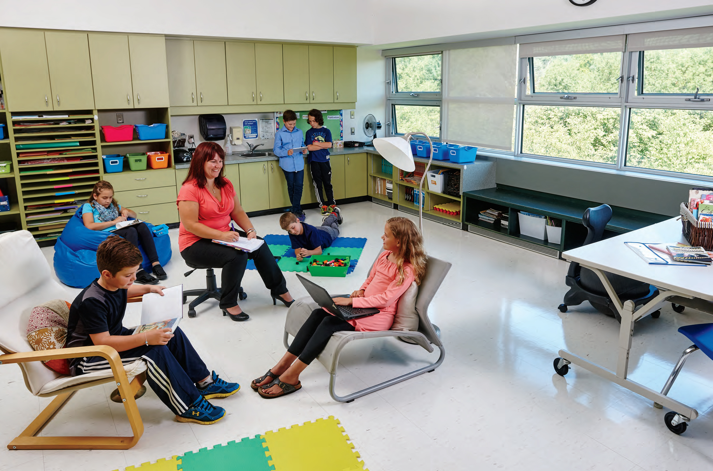 Mélissa Foisy, an Ontario Certified Teacher, is sitting in a classroom surrounded by young students. Mélissa is smiling and writing on a notepad. The students are engaged in various activities around the classroom.