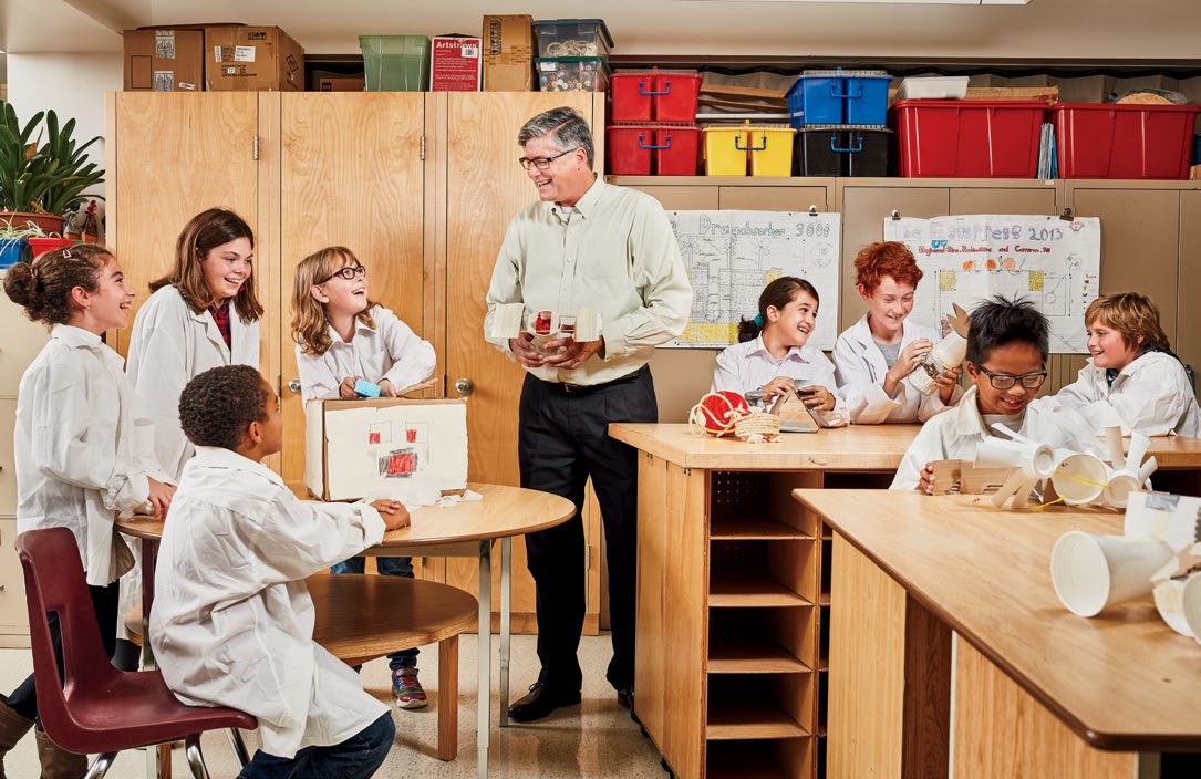 A photo of Steve Revington standing in a classroom. Steve is surrounded by young students wearing lab coats. The students are all working on various projects.