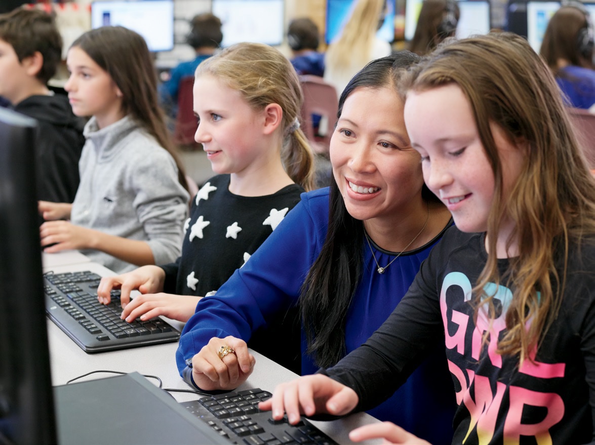 Photo of a computer lab filled with students working on computers. A teacher smiles as she crouches down to sit beside and help one student.