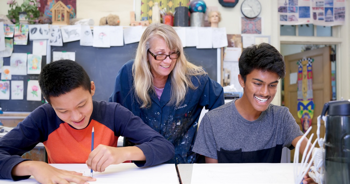 Photo of a teacher smiling while looking over the shoulder of two students who are working on an art task.