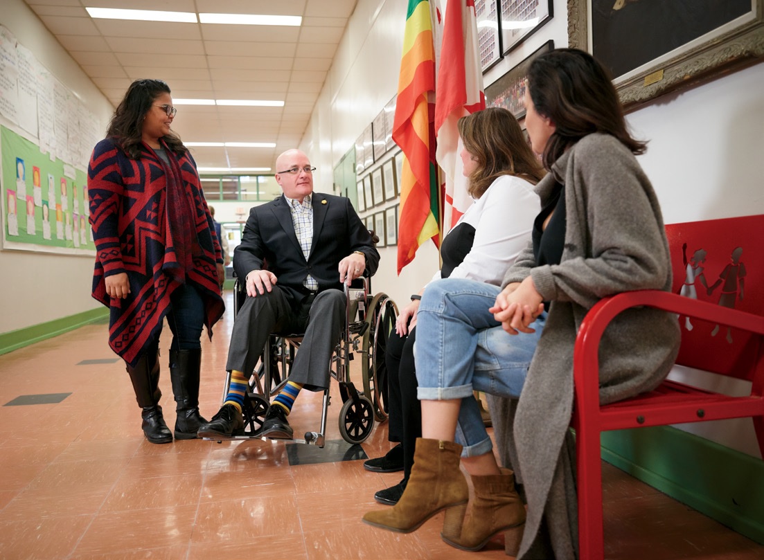 Photo of four people having a group discussion in the hallway of a school. One person is standing up beside a person in a wheelchair and there are two people sitting down on a bench.
