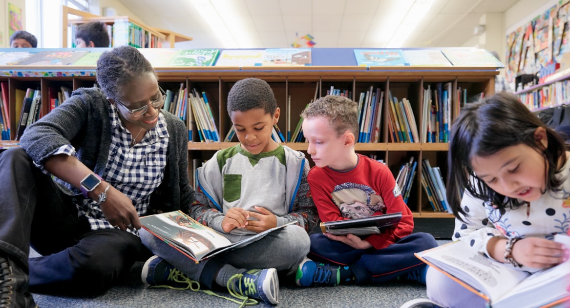 Photo of a teacher and students sitting on the floor in a library. The teacher is smiling while looking at a book with two students.