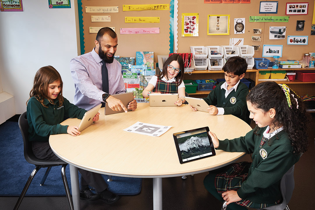 Photo of Afzal Shaikh, Ontario Certified Teacher, seated at a desk with a group of young students. They are all holding iPads.