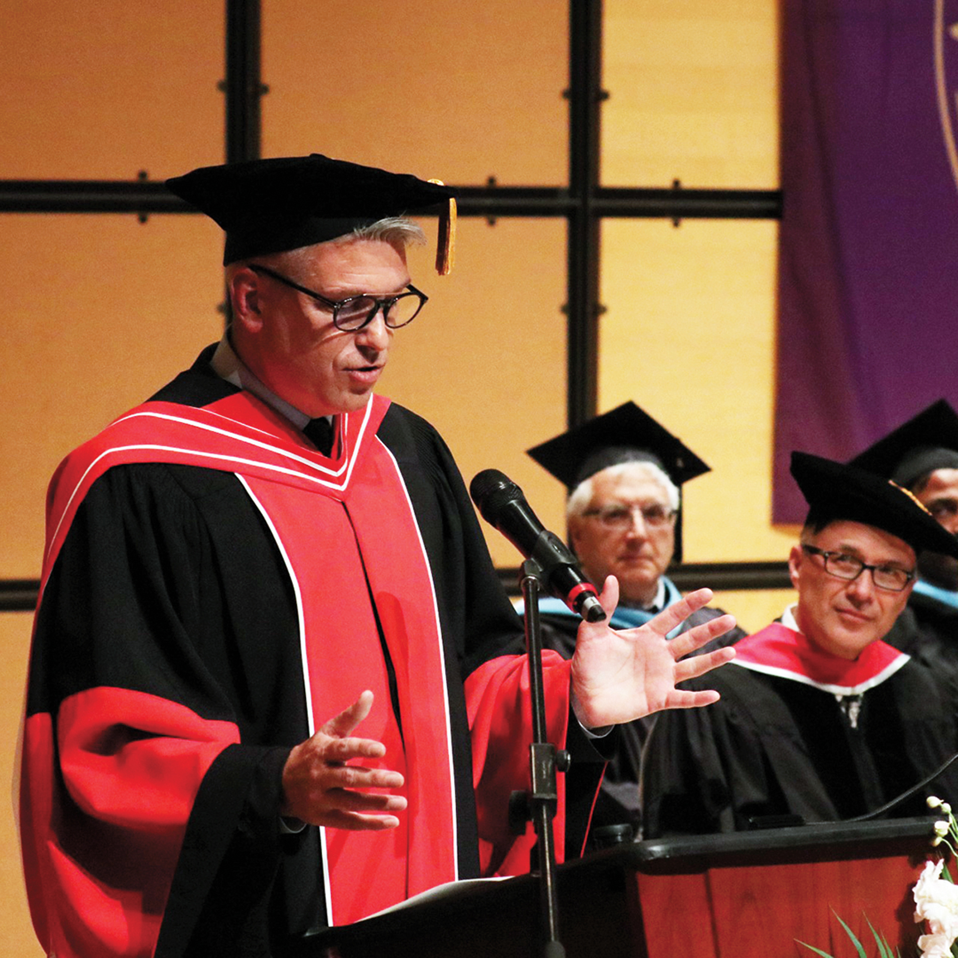 Photo of College CEO and Registrar Michael Salvatori wearing a commencement gown and cap while speaking into a microphone.
