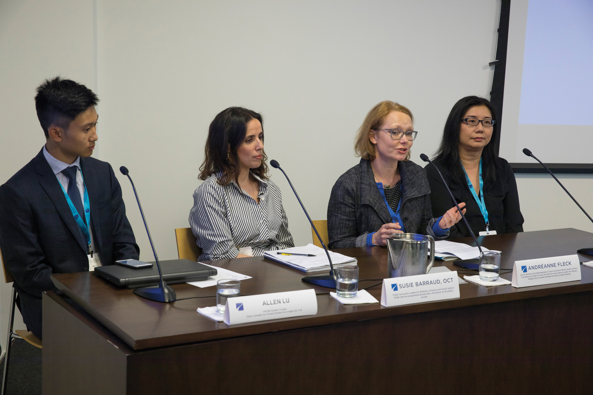 Photo of four people in a panel discussion, sitting at a desk, each in front of a microphone. One person is speaking into the microphone.