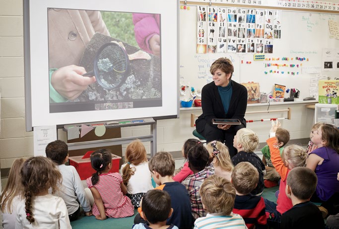 A photo of Angela Harrison seated in front of a whiteboard at the head of a classroom. There is an entire class of young students seated on the floor, watching the whiteboard.