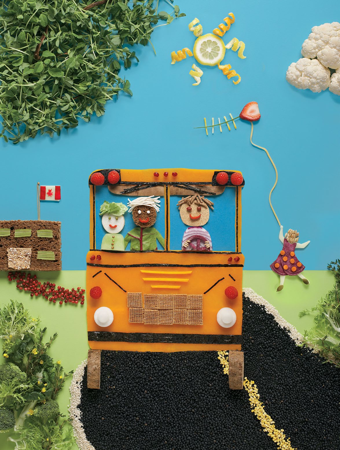 An image created out of various food items. There is a schoolbus crafted out of a slice of cheese driving on a road made of blueberries. Above the schoolbus is a lemon slice as the sun and cauliflower as clouds. In the foreground there is broccoli and mixed greens.