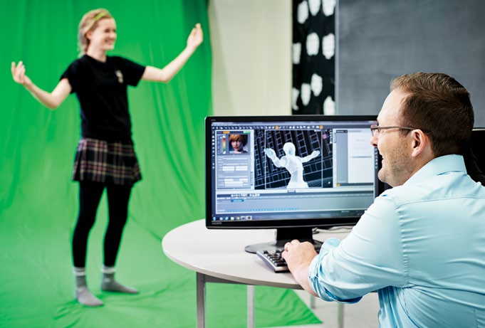 A photo of James Pedrech using a computer. In the background a student stands with arms wide. The student is in front of a green screen. James is creating a model of the student on the computer.