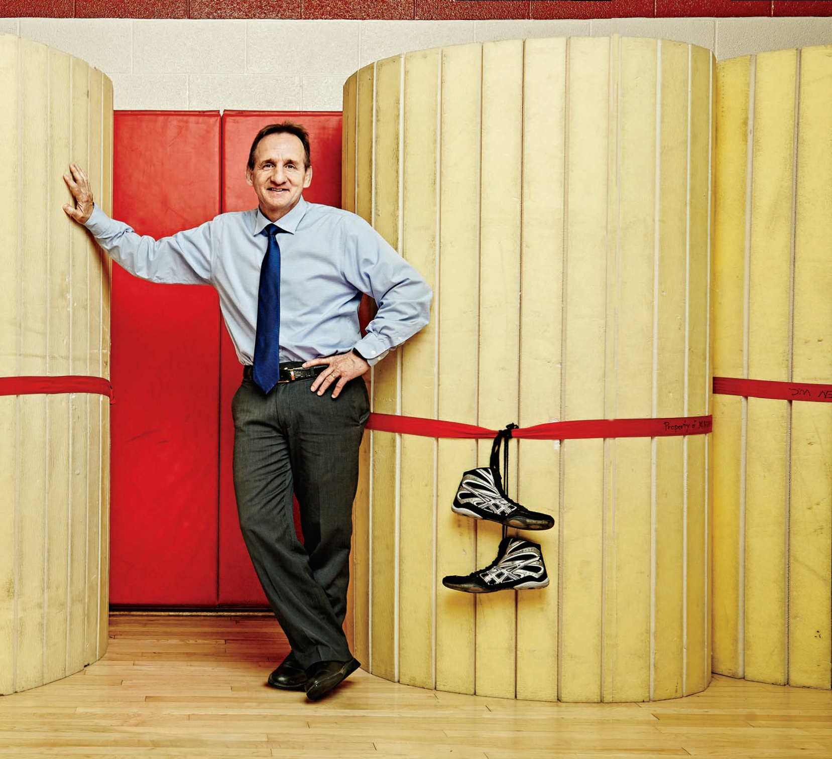 A photo of former Olympic wrestler, Chris Woodcroft, Ontario Certified Teacher, leaning on a wall. There are a pair of wrestling shoes tied to the wall next to Chris.