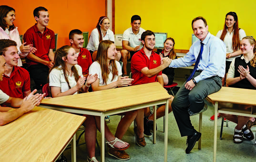 A photo of former Olympic wrestler, Chris Woodcroft, Ontario Certified Teacher, sitting on a desk surrounded by teenaged students. Chris is grasping hands with a student while the other students applaud.