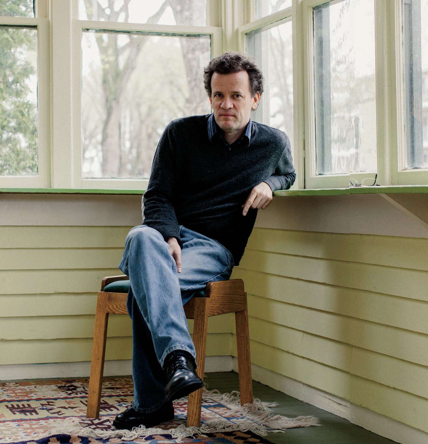 Photo of Bestselling author Yann Martel sitting on a stool and leaning on the windowsill. Yann is looking intently at the camera.