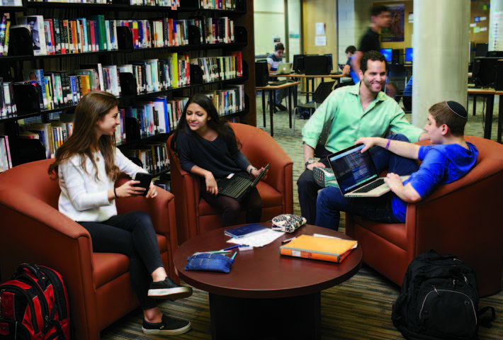 Photo of Jamie Cohen, Ontario Certified Teacher, seated with three students. One of the student is working on a laptop and talking with Jamie. The other two students are looking at a smartphone.