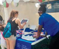 A representative of the Ontario College of Teachers leans over a table to assist a visitor using a touchscreen device at the Barrie Waterfront Festival.