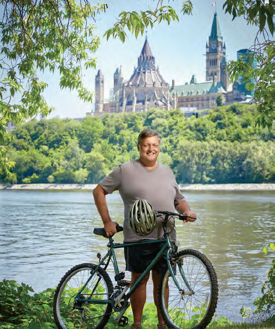 A photo of René Chiasson, retired Ontario teacher. René  is standing with a bike in front of the Rideau canal with Canada's Parliament in the background.