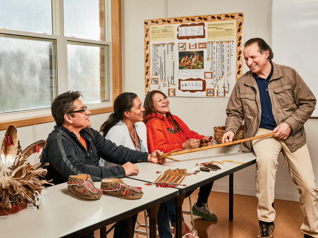 Photo of Tom Deer, Ontario Certified Teacher, seated on a table and pointing to a wooden implement. There are three students seated at the table watching Tom.