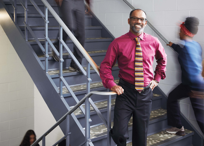 Photo of Michael Naicker, Ontario Certified Teacher, standing in a stairwell while students walk in the background.