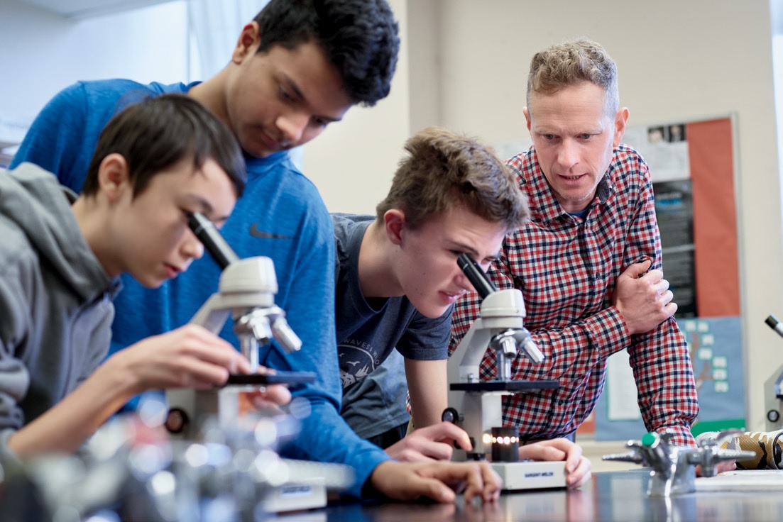 Photo of four people. Two are students looking, each looking through a microscope with a person standing to the right of them.
