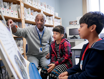 Photo of a teacher and two students sitting on chairs. The students observe as the teacher points to a section of a poster.