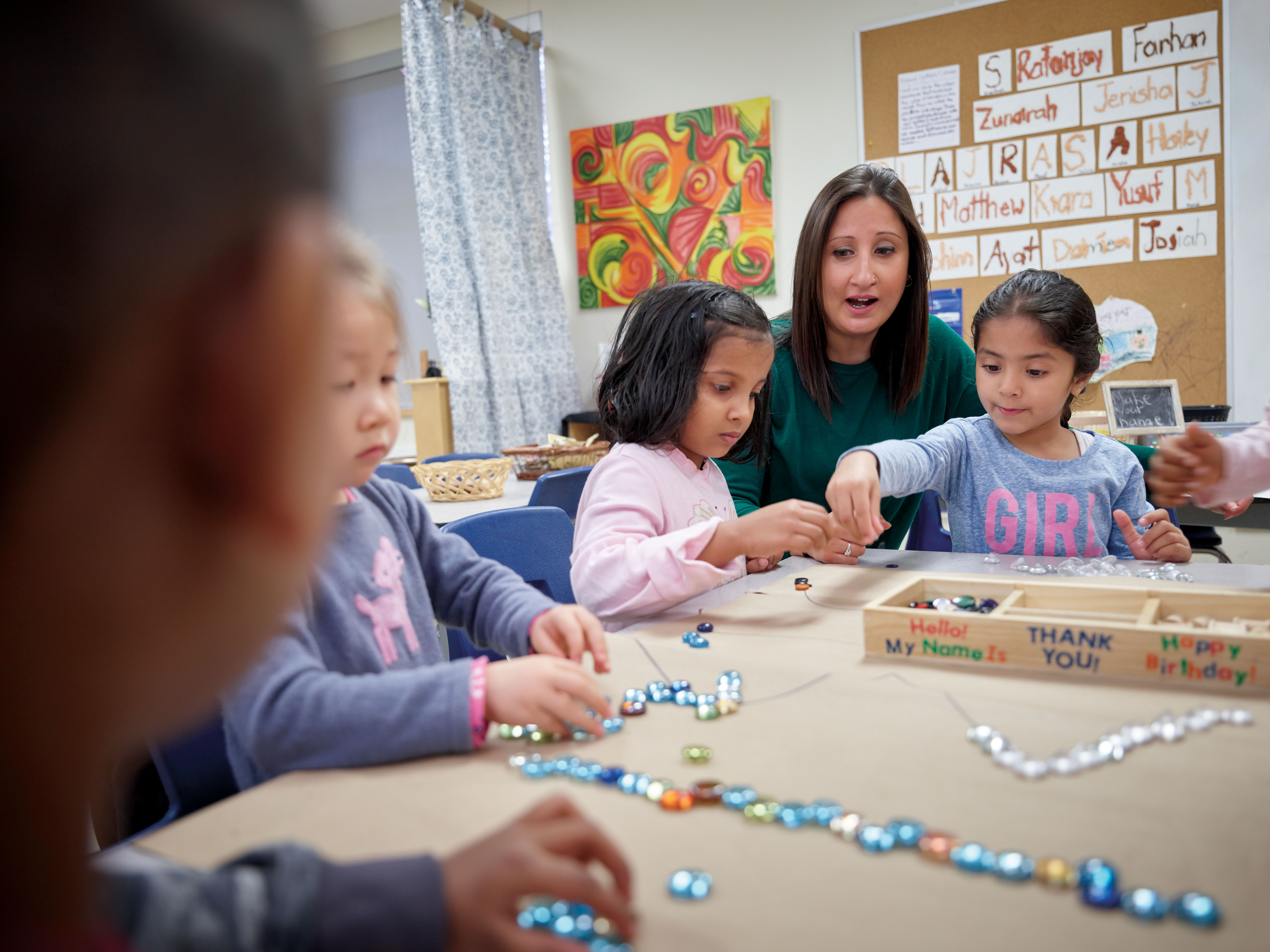 Photo of a teacher sitting down at a table with young students who are seated and picking up pieces of a project on the table.