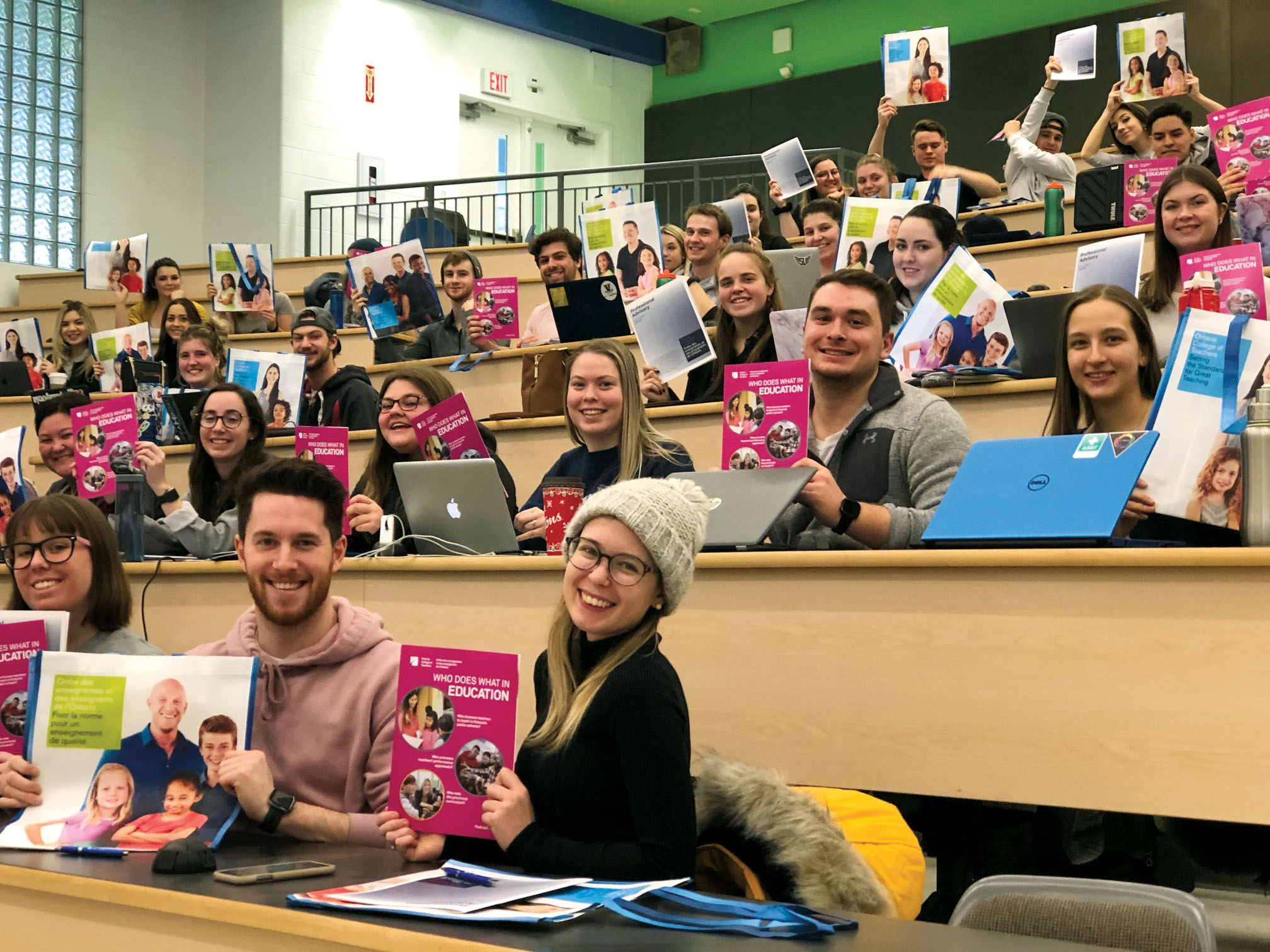 Photo of a large group of teacher candidates in a lecture hall, smiling and holding brochures and bags.